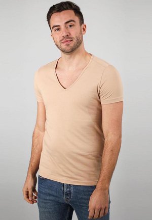 Man met kort donker haar en getrimde baard, gekleed in een beige V-hals T-shirt met korte mouwen en blauwe spijkerbroek tegen een effen grijze achtergrond.