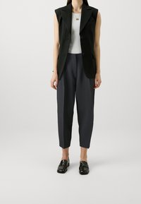 Blazer sans manches noir sur un haut côtelé blanc, associé à un pantalon gris foncé taille haute et fuselé et des mocassins noirs.