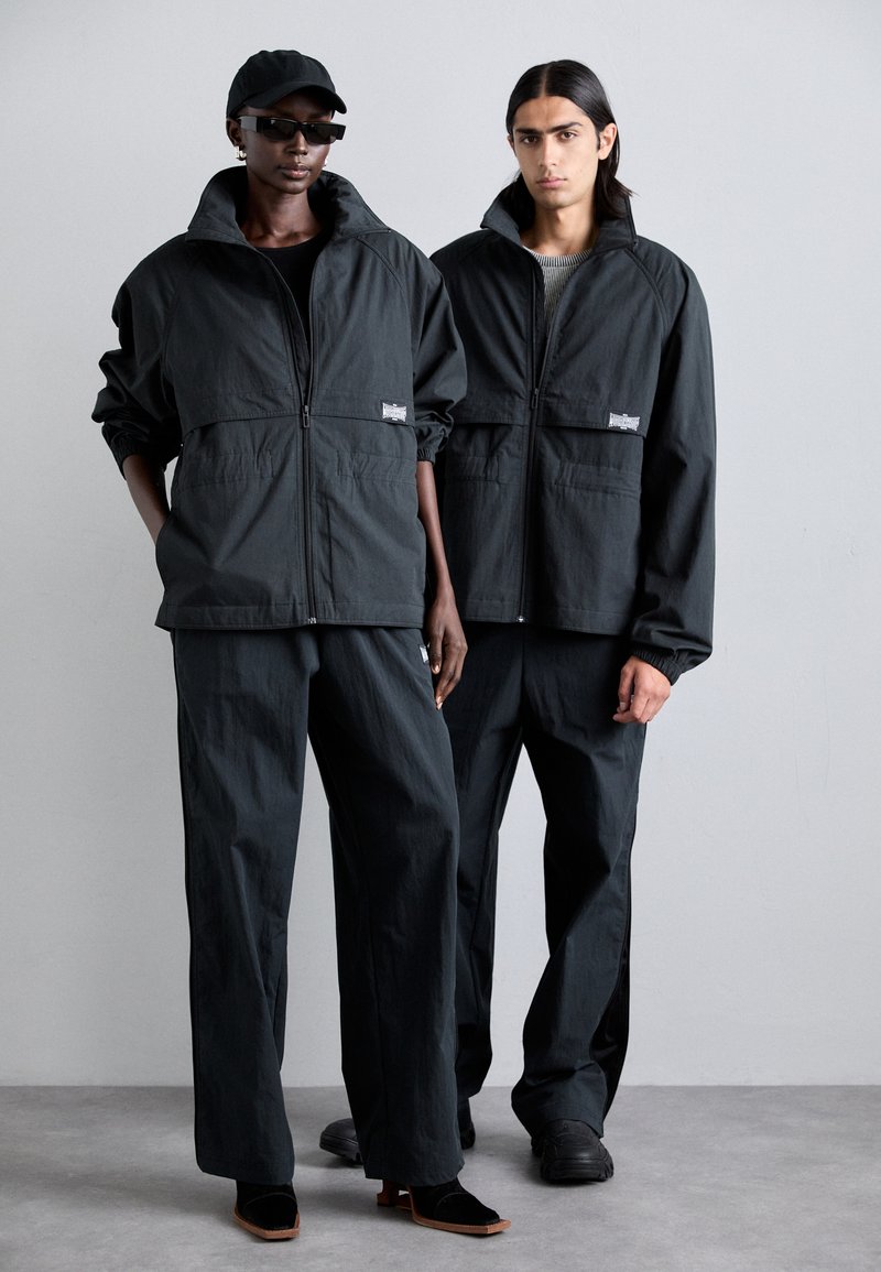 Conjunto exterior negro que incluye chaquetas con cremallera y cuellos alzados, combinadas con pantalones sueltos a juego. Los complementos incluyen una gorra y gafas de sol.