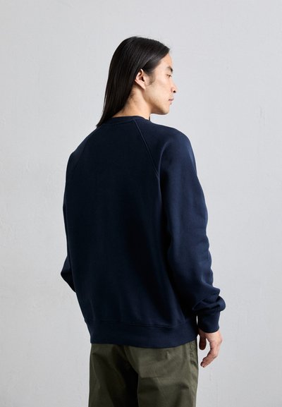 Sudadera azul marino de tejido suave, con cuello redondo acanalado, mangas raglán y un corte relajado, combinada con pantalones verde oliva.