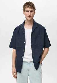Cămașă scurtă cu mânecă, de culoare bleumarin, cu un model texturat, două buzunare pe piept și butoane albe, asortată cu un tricou alb dedesubt.