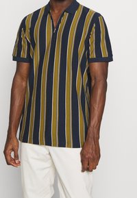 Kortärmad polo skjorta i marinblått, olivgrönt och vitt vertikalt randig. Har en dragkedjsstängning och ribbad krage. Mjuk bomullsmaterial.