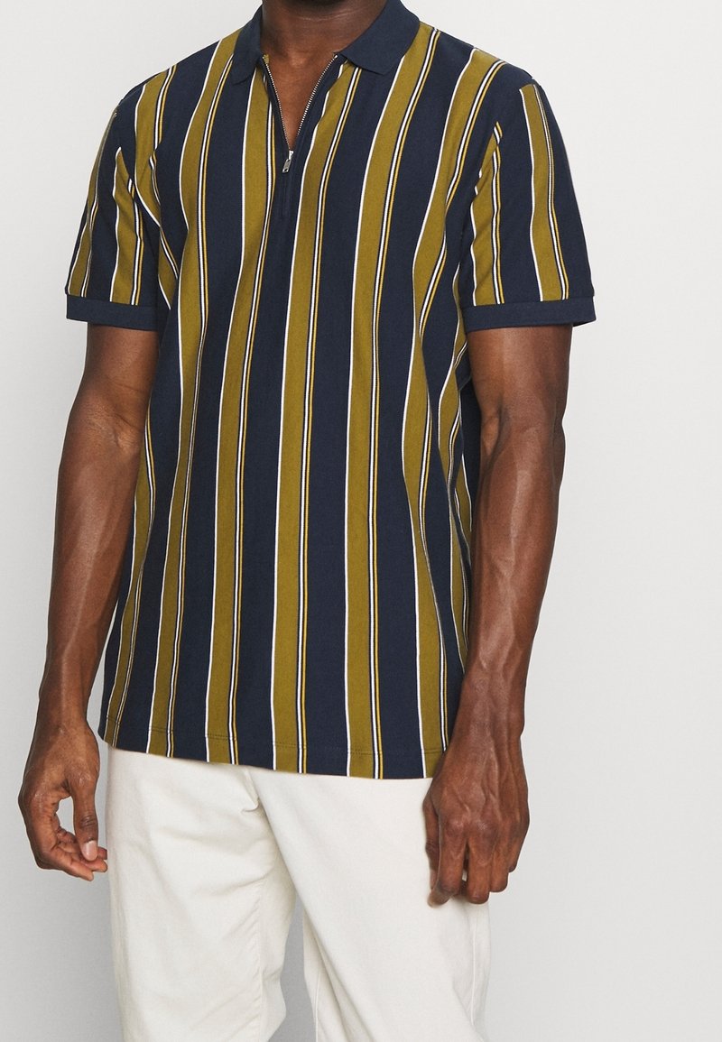 Kortärmad polo skjorta i marinblått, olivgrönt och vitt vertikalt randig. Har en dragkedjsstängning och ribbad krage. Mjuk bomullsmaterial.