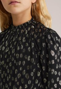 Crna odjeća s naboranim izrezom, koja sadrži metalik zlatne i srebrne uzorke životinja. Teksturirana tkanina, napuhane rukave i plisirani dizajn.