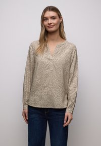 Blouse beige à manches longues avec un imprimé animalier discret, dotée d'un décolleté en V et d'une coupe décontractée. Portée avec un jean bleu.