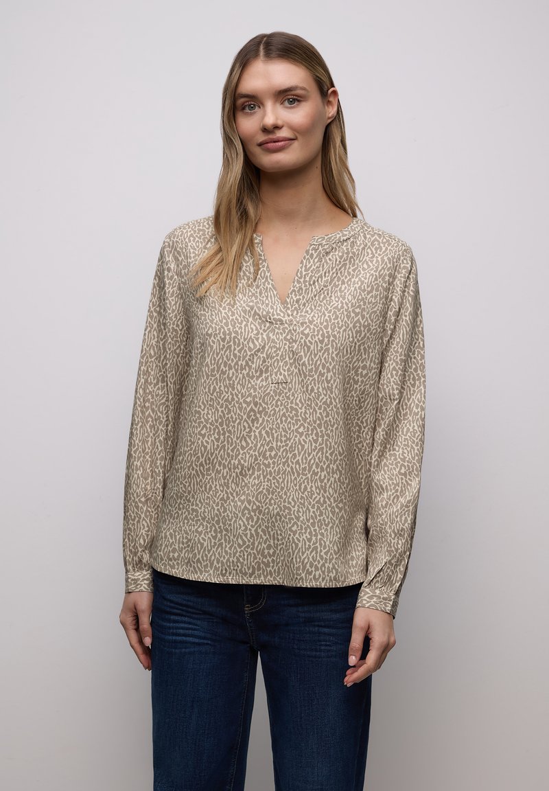 Blouse beige à manches longues avec un imprimé animalier discret, dotée d'un décolleté en V et d'une coupe décontractée. Portée avec un jean bleu.