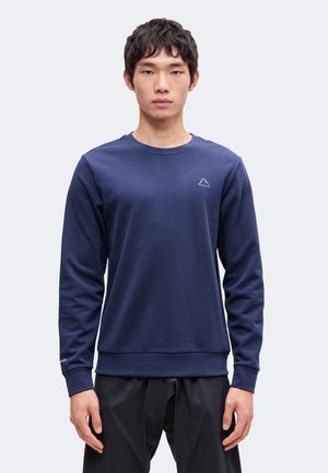 Jeune homme portant un sweat-shirt bleu marine uni avec un petit logo montagne sur la poitrine et un pantalon noir, debout devant un fond clair.