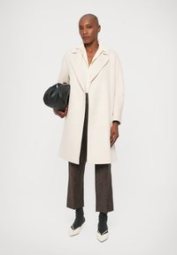 Cappotto di lana beige chiaro con ampi revers, abbinato a una camicia chiara e pantaloni marroni a fantasia. Tiene una borsa nera. Scarpe bianche a punta.
