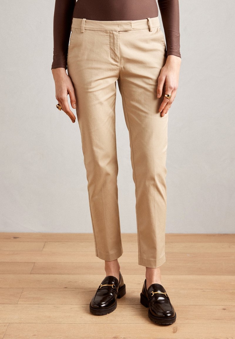 Personne portant un pantalon beige ajusté longueur cheville, un haut marron à manches longues, des bagues en or et des mocassins noirs, debout sur un sol en bois.