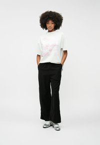 T-shirt en coton blanc avec un design graphique rose, associé à un pantalon noir à jambes larges et des chaussures argentées. Le modèle se tient avec les mains dans les poches.