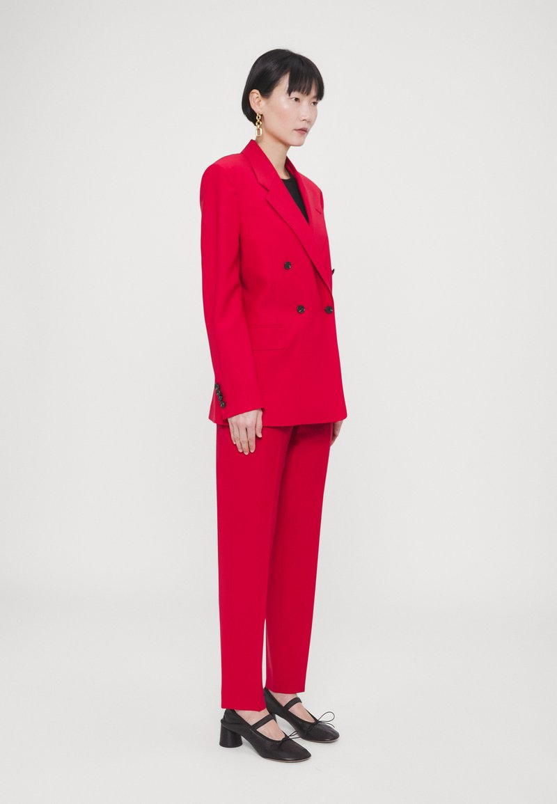 Blazer rouge vif à double boutonnage avec des boutons noirs, associé à un pantalon rouge assorti. Des chaussures noires à talons bas et des boucles d'oreilles en or complètent le look.