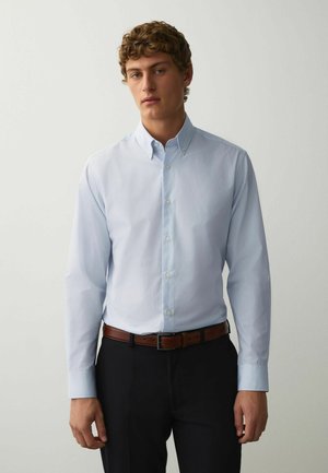 Junger Mann mit lockigem Haar, der ein hellblaues Hemd mit Knöpfen, dunkle Hosen und einen braunen Gürtel trägt, steht vor einem schlichten grauen Hintergrund.