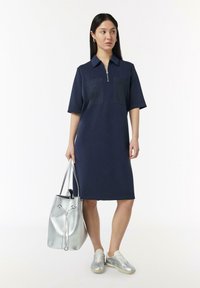 Marineblauwe jurk met korte mouwen, een ritssluiting bij de hals en twee borstzakken; gecombineerd met zilveren sneakers en een glanzende zilveren handtas.