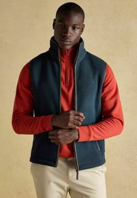 Tumši tīklots fleece bezrūpīgs veste uz sarkanas garām piedurknēm krekla, ar augstu apkaklīti un rāvējslēdzēju. Rokas sakrustotas priekšā, pret neitrālu fonu.