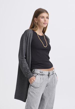 Dlhý sivý kardigán z mäkkej látky, otvorený vpredu, s dlhými rukávmi. Nosí sa cez priliehavý čierny top a sivé nohavice s vysokým pásom.