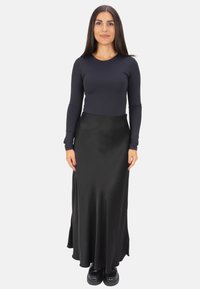 Frau mit langen dunklen Haaren, die ein enges schwarzes Langarmoberteil, einen schwarzen Satin-Maxirock und schwarze Plateauschuhe trägt, vor einem weißen Hintergrund steht.