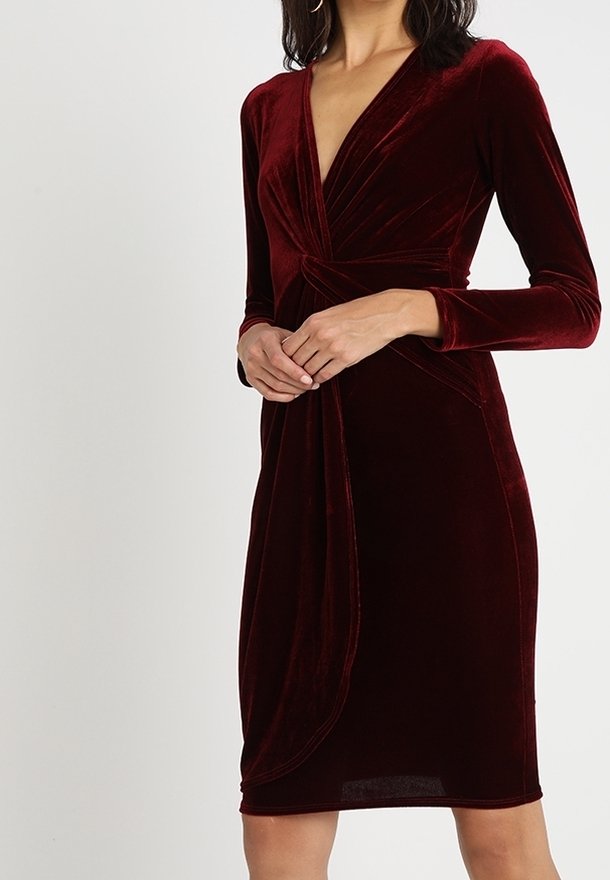 Femme portant une robe moulante en velours rouge profond à manches longues, col en V et détail froncé à la taille.