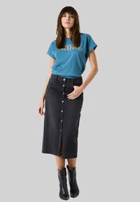 T-shirt bleu avec texte doré, associé à une jupe en denim noir avec un détail de bouton central et une fente avant, complété par des bottines noires.