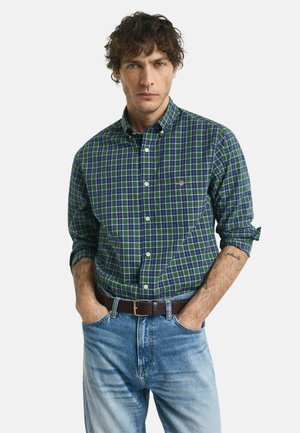 Mann mit lockigem Haar, der ein grün-blau kariertes Hemd trägt, das in eine hellblaue Jeans gesteckt ist, mit den Händen in den Taschen.