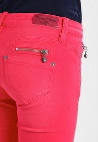 Pantalones rosa brillante con un bolsillo trasero, un bolsillo con cremallera para monedas, botones de metal y una etiqueta de marca de cuero marrón encima de la cinturilla.