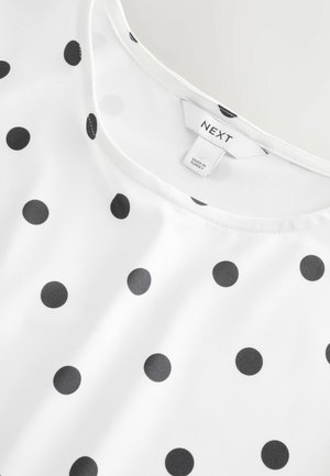 Hvid stofbluse med sorte prikker, rund halsudskæring og et mærke med teksten "NEXT." Glat tekstur med ensartet placering af prikkerne.