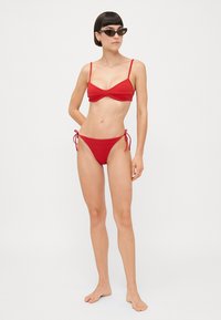 Červený bikiny set s jednoduchým braletovým topom s nastaviteľnými ramienkami a nízko strihanými bokovými nohavičkami na viazanie. Hladká textúra s minimálnym zdobením.