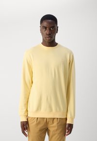 Gelbes langärmeliges Sweatshirt aus weichem Baumwollmaterial mit geripptem Kragen und Bündchen, das ein kleines Logo auf der Brust trägt. Trägt man über beigen Hosen.