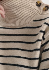 Suéter de punto beige con rayas horizontales negras, que presenta un alto cuello acanalado y dos botones redondos marrones en el hombro.