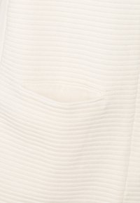 Nahaufnahme eines weißen gestrickten Stoffes mit horizontaler Rippstruktur und einem subtilen ovalen, erhabenen Muster in der Mitte.