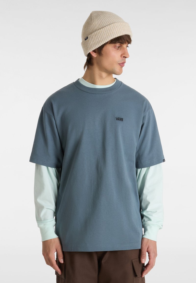 Camiseta azul de manga corta con cuello redondo y detalle del logotipo negro, superpuesta a una camisa de manga larga azul claro. (lleva un) gorro de punto beige en la cabeza.