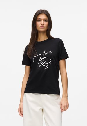 Mlada žena s dugom tamnom kosom nosi crnu majicu s bijelim rukopisnim tekstom "from Paris love, Karl xx" i bijele hlače, stojeći nasuprot bijeloj pozadini.