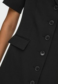 Crna krojena haljina s kratkim rukavima, koja sadrži pet velikih crnih dugmadi i detalj s preklopnom džepom na lijevoj strani. Teksturirana tkanina.