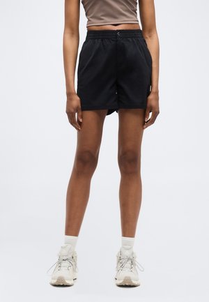 Persona que lleva pantalones cortos negros, top corto beige, calcetines blancos y zapatillas deportivas blancas, de pie contra un fondo liso.