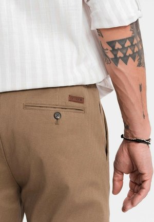 Brune bukser lavet af struktureret stof, med en baglomme med en knap og et mærket lædermærke. Skjorten er hvid med fine striber.