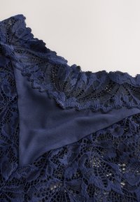 Tessuto in pizzo blu navy con bordo smerlato, sovrapposto a un materiale satinato liscio, caratterizzato da intricati motivi floreali e dettagli strutturati.