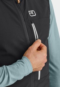 Schwarze Weste mit weißem Reißverschluss und Logo-Detail, aus strapazierfähigem Material gefertigt. Der Reißverschluss lässt sich mit der Hand verstellen und zeigt eine Tasche auf der Vorderseite. Lange blaue Ärmel sind sichtbar.