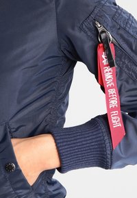 Mörkblå bomberjacka med en glänsande textur, ribbad mudd och en röd tyglabel som står "REMOVE BEFORE FLIGHT" fäst vid dragkedjan.