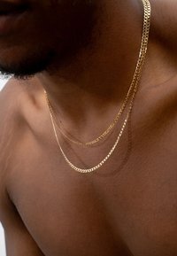 Goldfarbene Metallketten mit flachen und gedrehten Gliedern, in drei Strängen geschichtet, ruhend auf brauner Haut. Glatte Textur und glänzende Oberfläche.