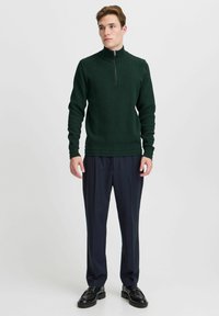 Dunkelgrüner gerippter Pullover mit hohem Kragen und halbem Reißverschluss. Kombiniert mit marineblauen plissee Hosen und schwarzen Lederschuhen.