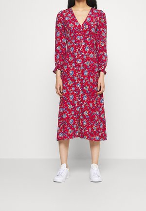 Mujer con vestido midi rojo con estampado floral azul, mangas largas y zapatillas blancas, de pie frente a un fondo claro y liso.