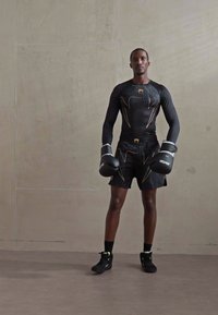 Un athlète portant des gants de boxe noirs, un tee-shirt de compression à manches longues et un short, se tenant contre un mur gris texturé.