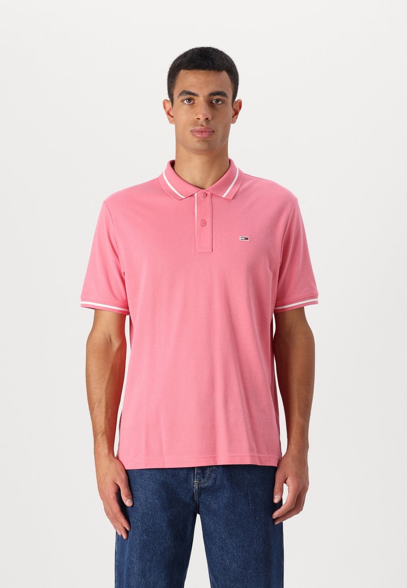 Rosa polo skjorte med en knappelukking med to knapper, hvit kant på kragen og ermene, med et lite logo på brystet. Myk stofftekstur.