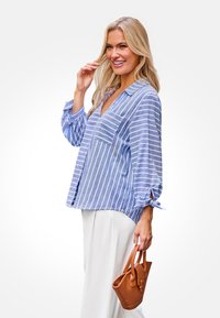 Hellbluse mit Streifen, V-Ausschnitt, langen Puffärmlen, die an den Bündchen gebunden sind, und einer Brusttasche, kombiniert mit cremefarbenen Hosen und einer braunen Handtasche.