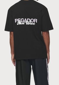 Černé tričko s bílým a růžovým textem na zádech, které zní "PEGADOR Next Wave." Krátké rukávy a uvolněný střih.