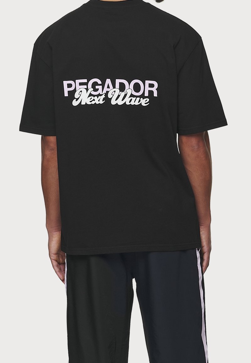 Černé tričko s bílým a růžovým textem na zádech, které zní "PEGADOR Next Wave." Krátké rukávy a uvolněný střih.