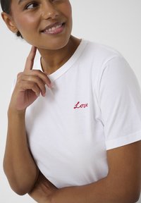 Personne souriante avec une main touchant le visage, portant un t-shirt blanc brodé du mot "Love" en rouge sur la poitrine.