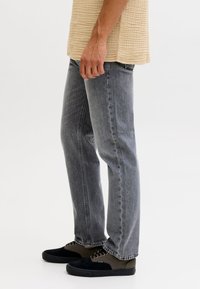 Mees seisab, kandes tuhmiks kulunud musti teksaseid, beeži topi ja mustade ning oliiviroheliste paeltega tossusid valgel taustal.