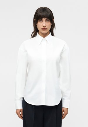 Žena s tmavými vlasy nosí bílou košili s knoflíky, špičatým límcem a dlouhými rukávy, v kombinaci s tmavými kalhotami, stojící před jednobarevným pozadím.