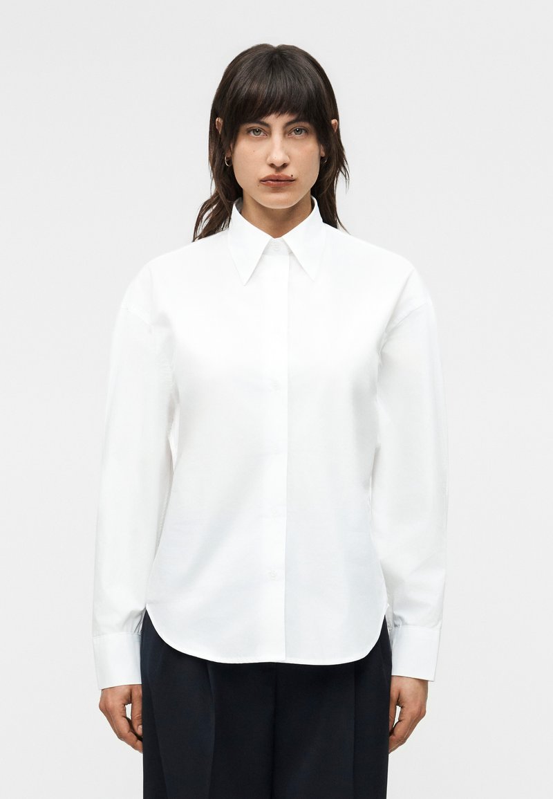 Tume juustega naine kannab valget nööpidega särki, millel on terava kraega ja pikad varrukad, koos tumedate pükstega, seistes ühevärvilise tausta ees.