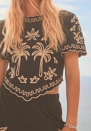 Sort cropped top med beige broderede palmetræer, blade og bølget trim, båret med matchende sorte bukser og et sølvkædearmbånd.